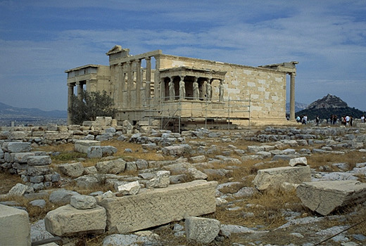 General view of the Erechtheion from the southwest, showing the North Porch (at left), the Olive Tree of Athena (modern replacement!), and the Carytatid Porch. - Note the modern (white) blocks of Pentelic marble which have been used in the reconstruction of the building. The blue limestone foundations of the Old Athena Temple (built c. 510-500 BC and destroyed by the Persians in 480 BC) are visible south of the E
